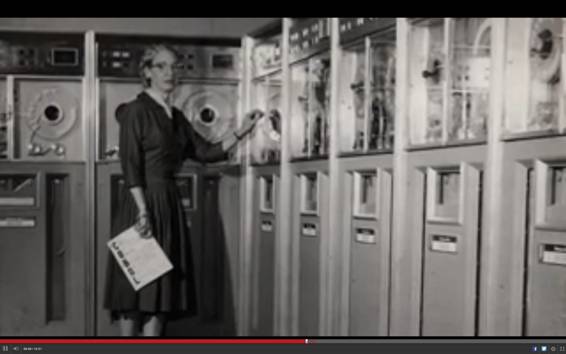 Grace Hopper ante unas cuantas unidades de almacenamiento de la época con un libro sobre COBOL en las manos